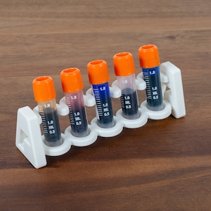 Puede incluir: Seis pequeños viales de vidrio transparente con tapas naranjas y líquido negro en su interior. Los viales están en un soporte de plástico blanco con los números 1,0 y 0,5 impresos en el lateral.