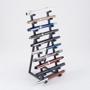 May include: A black metal pen display stand showcasing ten fountain pens. The pens feature diverse designs and colours, including gold, blue, and wood-effect finishes. The stand is angled for display, offering a clear view of each pen.