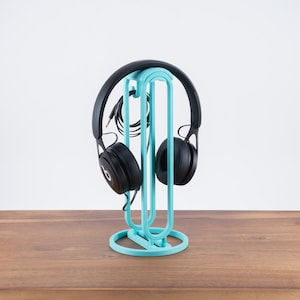 Può includere: Un supporto per cuffie di colore blu turchese con due cuffie nere appese. Il supporto ha una base circolare.