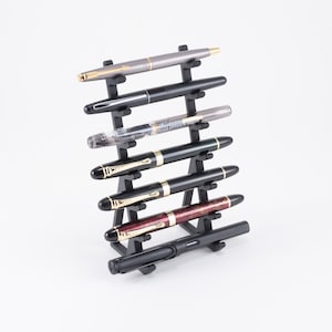 May include: A black pen stand with seven pens, including a black and gold pen, a red and gold pen, and a black pen with a silver clip.