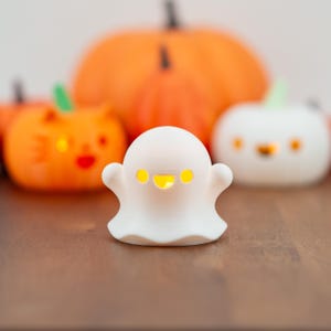 May include: A white ghost-shaped Halloween decoration with a smiling face and glowing yellow eyes. In the background are orange pumpkin decorations, creating a festive autumn scene. The ghost is on a wooden surface.
