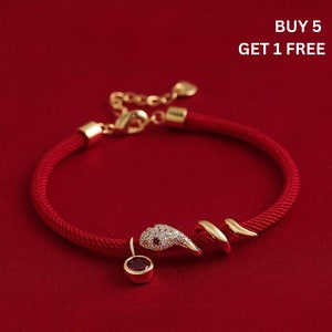 May include: Red rope bracelet featuring a gold-coloured snake design and a small, round, red charm. The snake's head is encrusted with small, clear stones. The bracelet is set against a red background. Text in the image reads: BUY 5 GET 1 FREE.