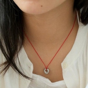 May include: A woman wearing a white cardigan with a red string necklace and a small silver heart pendant.