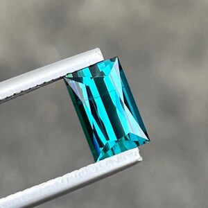 以下が含まれることがあります： ピンセットで保持された長方形のターコイズブルーの宝石。宝石はストライプ模様で光沢のある仕上げになっています。
