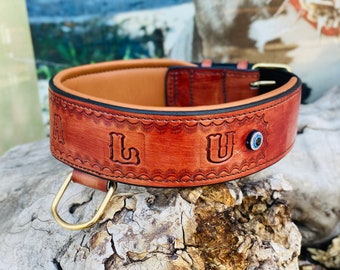 Puede incluir: Un collar de perro de cuero marrón con las letras "LU" grabadas. El collar tiene una hebilla de metal dorado y un amuleto azul de ojo turco.