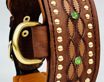 May include: A brown leather dog collar with gold studs and green gemstones. The collar has a decorative leaf pattern and a brass buckle.