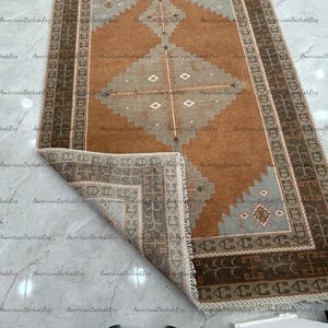 May include: A rectangular area rug featuring a central diamond motif in light grey against a warm brown field. The rug has a dark brown border with geometric patterns and a light-coloured fringe. One corner is folded back to reveal the underside.