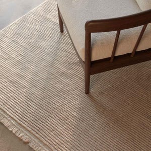 Könnte beinhalten: Ein beigefarbener, strukturierter Teppich mit linearem Muster und Fransenrand. Ein Holzrahmen-Sessel mit cremefarbenem Kissen steht auf dem Teppich. Die neutrale Farbe und Textur des Teppichs deuten auf Inneneinrichtung hin.