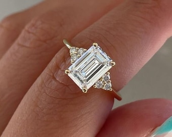 May include: A gold ring featuring an emerald-cut diamond center stone, flanked by smaller accent diamonds. The ring's band is a delicate gold, and the setting showcases the diamonds' brilliance. This elegant piece is a classic choice for engagement or special occasions.