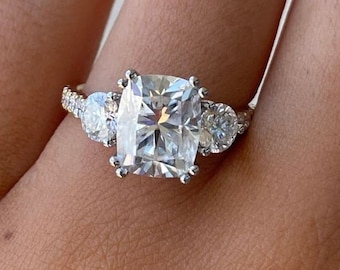 May include: A three-stone engagement ring with a large, square-shaped center stone and two smaller round stones on either side. The ring is set in white gold and has a pave setting.