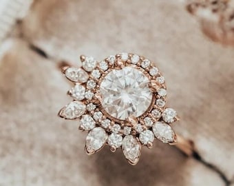 May include: A rose gold engagement ring with a large round center stone surrounded by smaller diamonds. The ring is set in a halo style with a delicate floral design.