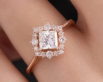 May include: A rose gold engagement ring with a square-shaped center stone surrounded by smaller diamonds. The ring has a delicate, intricate design with a halo of smaller diamonds around the center stone.
