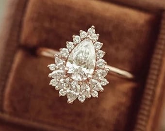 May include: A gold ring with a pear-shaped diamond center stone surrounded by smaller diamonds. The ring is set in a brown velvet ring box. The ring's design is elegant and suitable for special occasions.