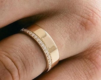 May include: A gold ring with a flat band and a row of small, clear gemstones set along one edge. The ring is worn on a finger, and the metal has a polished finish. The ring is a piece of jewelry.
