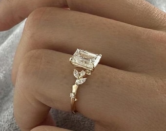 May include: A gold ring with an emerald-cut diamond center stone. The ring features a delicate leaf design with smaller accent diamonds. The ring is shown on a hand against a soft gray background.