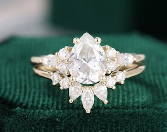 May include: A pear-shaped diamond engagement ring with a halo of smaller diamonds set in a gold band. The ring is on a green velvet surface.