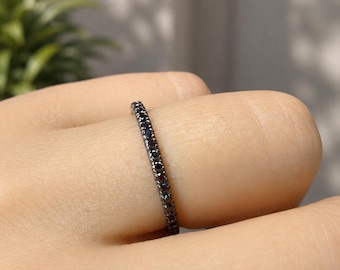 May include: A thin, dark metal band ring featuring a continuous row of small, round, dark-colored gemstones set along the outer surface. The ring has a slim profile and a polished finish, showcasing a series of uniform, dark, sparkling stones.