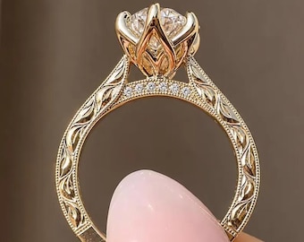 May include: A gold ring featuring a large round brilliant-cut center stone held in a tulip-shaped setting. The band is decorated with intricate leaf-like engravings and a row of small pavé-set diamonds along the upper shank.