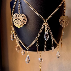 May include: A gold-toned metal chain plant hanger featuring dangling metallic monstera leaf charms and clear, faceted teardrop-shaped crystals. The decorative chain assembly is draped around a matte black cylindrical plant pot.