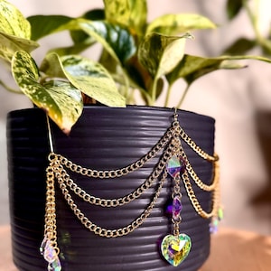 May include: A black, ribbed ceramic plant pot with a trailing golden chain and crystal embellishments. The chains hang in layers, adorned with iridescent crystals and a heart-shaped charm. The plant has green and yellow leaves.
