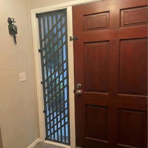 Puede incluir: Una puerta de seguridad de metal gris oscuro con un patrón de diamantes está instalada junto a una puerta de madera marrón oscuro. La puerta de seguridad está enmarcada en blanco y tiene un asa. La puerta de madera tiene un pomo plateado.