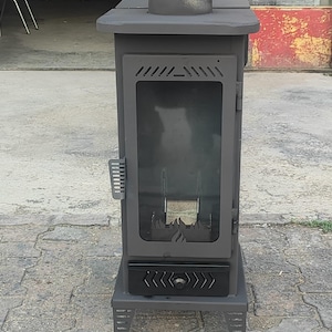 Puede incluir: Una estufa de leña de metal gris carbón con una puerta de cristal rectangular que presenta un recorte de metal en forma de llama en la base, un collarín de humos circular superior, un asa lateral y un cajón inferior con pomo central.