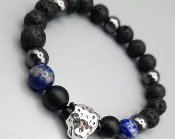 May include: A beaded bracelet featuring black lava rock beads, matte black beads, silver-colored hematite beads, and blue lapis lazuli beads. The bracelet includes a silver-colored, textured focal bead. The bracelet is on a light gray background.