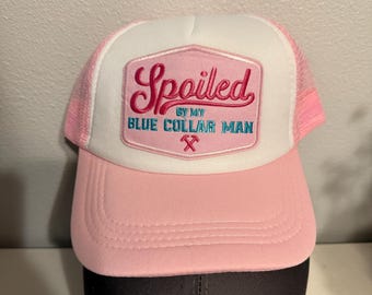 Può includere: Cappellino da camionista rosa e bianco con una toppa rosa che dice "Viziato dal mio uomo colletto blu" con un martello e una chiave inglese incrociati.