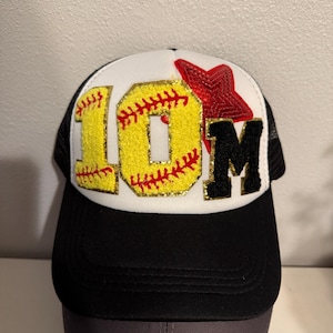 May include: Black and white trucker hat with a white front panel. The front panel features a red star, a yellow softball with red stitching, and the letters "10M" in black and yellow.