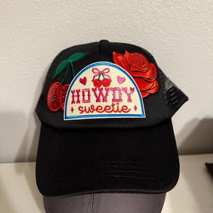 Puede incluir: Gorra de camionero negra con un parche blanco que dice "HOWDY sweetie" con cerezas y una rosa roja bordadas.