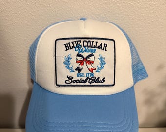 Può includere: Un cappello da camionista blu e bianco con una toppa che dice "Blue Collar Wives Est. 1776 Social Club" con un fiocco rosso, bianco e blu.