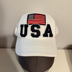 Może przedstawiać: Biała czapka bejsbolówka z naszywką amerykańskiej flagi w kolorze czerwonym, białym i niebieskim oraz napisem "USA" w niebieskich literach.