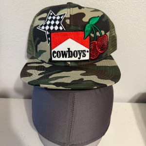 Puede incluir: Una gorra de camionero de camuflaje con un parche rojo y blanco que dice "cowboys" y un parche de estrella blanco y negro. También hay un parche de cereza roja con tallos verdes.