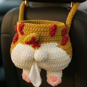 May include: A handmade, crochet tissue box cover shaped like a dog's rear end. The cover is mustard yellow with red spots, white fur, and pink paw pads. A tissue is pulled from the dog's rear. The cover hangs from a car headrest.