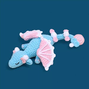 Peut inclure: Un jouet en crochet en forme de dragon bleu et rose avec des accents blancs. Le dragon a une queue enroulée et une petite bouche fermée.