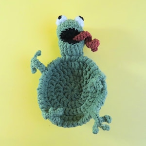 May include: Handmade crochet frog in shades of green, with white and black eyes, a wide open black mouth, and a red tongue. The frog has outstretched arms and legs, and is set against a yellow background. A whimsical, handmade item.