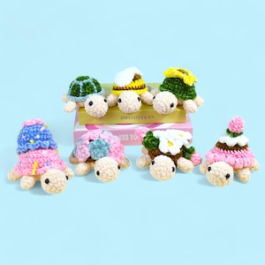 May include: Seven crocheted turtle toys with different colored shells and decorations. The turtles are arranged in a row on a pink and white box. The box has the text "LA PATISSERIE" on it.