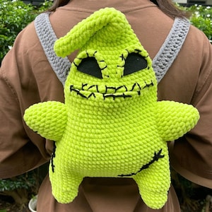 May include: A lime green crochet backpack shaped like a cartoon character with a stitched face and black accents. The backpack has a pointed top and attached gray straps.