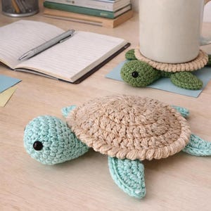 May include: Handmade crochet turtle coasters in shades of green, tan, and blue. The coasters are placed on a light-colored wooden surface, with a notebook, pen, and a mug in the background. The turtle coasters are a charming addition to any home.