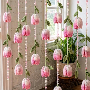 May include: Crocheted floral curtain with pink and white tulip-shaped flowers, green leaves, and beaded strands. The handmade curtain hangs from a wooden rod, adding a touch of spring to a room. The flowers are approximately 4 inches long.