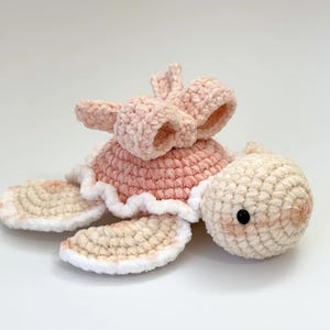 Puede incluir: Un juguete de tortuga de crochet con un lazo rosa en la espalda. La tortuga está hecha de hilo suave y de felpa y tiene un borde blanco alrededor de su caparazón y aletas.