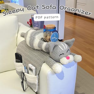 May include: A gray and white crocheted cat sofa organizer with pockets. The cat is lying down with its eyes closed and a pink nose. The organizer has pockets for a phone, glasses, and a remote control. The text "Sleepy Cat Sofa Organizer" and "PDF pattern" are visible.