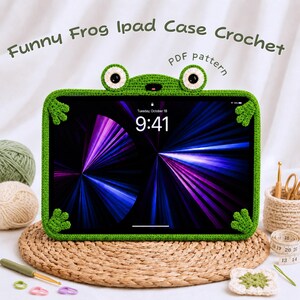 May include: A green crochet tablet case shaped like a frog with two eyes on top and four small frog feet at the corners. The case holds a tablet displaying a purple and blue light pattern. Text reads: Funny Frog Ipad Case Crochet PDF pattern.