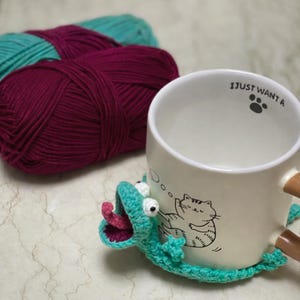 Puede incluir: Una taza de cer&aacute;mica blanca con una ilustraci&oacute;n de gato y el texto "I JUST WANT A" se encuentra sobre un posavasos de ganchillo turquesa con forma de lagarto. Dos madejas de hilo, una turquesa y otra burdeos, est&aacute;n en el fondo.