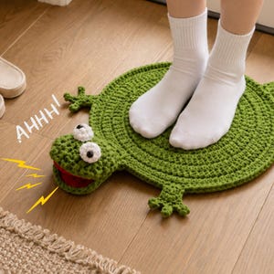 Può includere: Un tappeto rotondo all'uncinetto verde a forma di rana con una bocca larga e aperta, occhi bianchi con pupille nere e quattro piccole zampe. Il testo AHHH! è stampato in bianco vicino alla testa, accompagnato da grafiche di fulmini gialli.