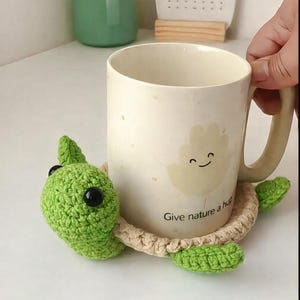 May include: A cream-colored mug with a smiling cloud design, resting on a handmade green and tan crochet turtle coaster. The coaster has a turtle head and flippers. The mug has the text "Give nature a hug".