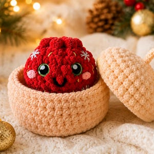 Peut inclure: Un contenant crochet&eacute; rond de couleur p&ecirc;che avec un couvercle assorti est pos&eacute; &agrave; c&ocirc;t&eacute; d'un petit personnage crochet&eacute; sph&eacute;rique rouge. Le personnage rouge pr&eacute;sente de grands yeux noirs, un sourire brod&eacute;, des joues roses et des flocons blancs.