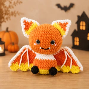 May include: A plush crochet bat toy featuring a color scheme of orange, white, and yellow, resembling candy corn. The bat has large black safety eyes, small pink cheek accents, two tiny white fangs, and small black feet at the base of its body.