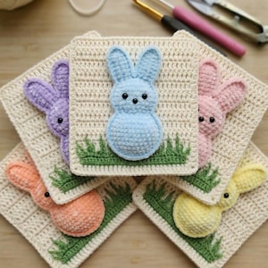 May include: A collection of five crocheted Easter bunny squares in pastel colors: blue, purple, pink, orange, and yellow. Each bunny is attached to a cream-colored square with green grass detail. Crochet hooks and scissors are in the background.