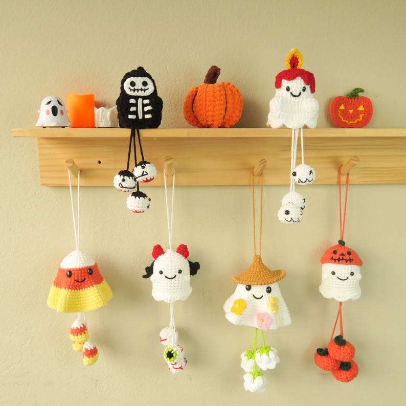 Op de afbeelding: Een verzameling handgemaakte gehaakte Halloween-ornamenten, waaronder een skelet, pompoen, spook en snoepma&iuml;s. De ornamenten zijn wit, oranje, geel en zwart en hangen aan een houten plank. De plank bevat ook een klein spookje, een kaars en een pompoen.